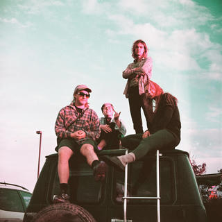 a film photo of four people sitting on top of a van, looking down at the camera. the film stock a purple tint, with heavy cyan as well